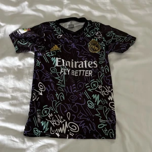 Real Madrid Vini Jr. Adidas fotbollströja - Snygg Real Madrid fotbollströja från Adidas med Vini Jr. och nummer 20 på ryggen. Tröjan är svart med färgstarka graffiti-inspirerade mönster i blått, vitt och lila. Tillverkad i lätt och ventilerande ClimaCool-material, perfekt för match eller träning.