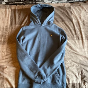 Blå hoodie från Lyle & Scott - Snygg blå hoodie från Lyle & Scott med klassisk gul logga på bröstet. Tröjan har huva, känguruficka och ribbade muddar. Perfekt för dig som gillar en stilren och bekväm look.