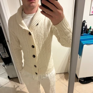 Vit kabelstickad kofta från Buddha - Säljer en vit kabelstickad kofta från Buddha med grov sjalkrage och stora bruna knappar framtill. Koftan har långärmad passform och är gjord i mjukt stickat material som känns riktigt chill. Perfekt för att hålla värmen under kyliga dagar. Bra skick, lätt nopprig. Storlek S men passar även xs 