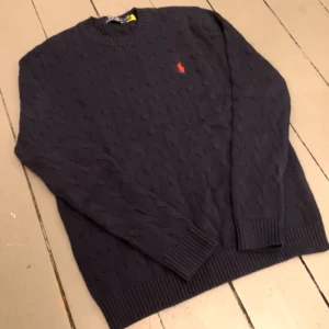 Mörkblå stickad tröja Polo Ralph Lauren - Mörkblå stickad tröja från Polo Ralph Lauren med klassiskt kabelstickat mönster och röd broderad logga på bröstet. Rund halsringning och ribbade muddar vid ärmslut och nederkant. Perfekt för dig som gillar stilrena och tidlösa plagg.