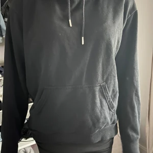 Svart hoodie med huva och ficka - Svart hoodie med klassisk huva och snörning, samt en stor magficka framtill. Tröjan har långärmade ärmar och en loose passform som gör den chill och enkel att bära. Perfekt för dig som gillar enkel och stilren streetwear.