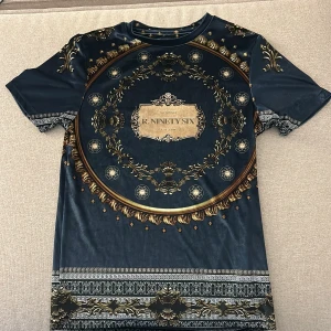 Svart och guld muscle fit t-shirt River Island - Unik muscle fit t-shirt från R-Ninety Six i svart med detaljerat barockinspirerat mönster i guld. T-shirten har korta ärmar och ett lyxigt tryck både fram och bak, med dekorativa kanter och inslag av silver. Perfekt för dig som gillar att sticka ut.