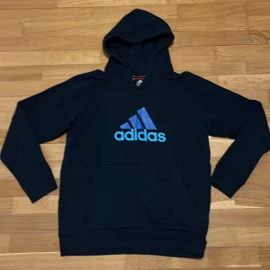 Mörkblå Adidas hoodie med logga - Snygg mörkblå hoodie från Adidas med stor blå logga på bröstet. Tröjan har huva och en praktisk magficka framtill. Perfekt för chill eller träning, med långärmade ärmar och mjukt material som känns skönt mot huden.