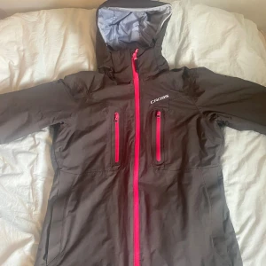 Brun skidjacka med rosa dragkedjor Cross - Snygg brun vindjacka från Cross med huva och rosa dragkedjor framtill. Jackan har två bröstfickor med dragkedja och är tillverkad i ett slitstarkt syntetmaterial. Perfekt för blåsiga dagar och outdoor-aktiviteter.