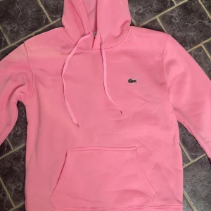 Rosa hoodie från Lacoste med huva - Säljer en snygg rosa hoodie från Lacoste i regular fit. Tröjan har huva med snörning, klassisk känguruficka och den ikoniska krokodilloggan broderad på bröstet. Perfekt för dig som gillar stilrena och bekväma plagg.