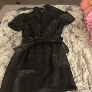 Svart kort klänning i skinnimitation - Snygg svart kort klänning i skinnimitation med knappar framtill och tillhörande bälte i midjan. Klänningen har korta ärmar och klassisk krage, perfekt för en edgy look.