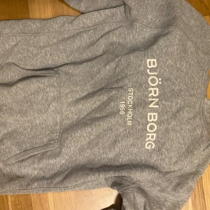 Grå hoodie från Björn Borg - Snygg grå hoodie från Björn Borg med tryck på bröstet där det står 'BJÖRN BORG STOCKHOLM 1956'. Tröjan har en klassisk känguruficka framtill och mjukt material som känns skönt mot huden. Perfekt för chill dagar eller när du vill ha en sportig look.