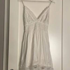 Vit spetsklänning med smala axelband - Supersöt vit kortklänning med smala axelband och v-ringad spets upptill. Klänningen har smockad midja och spetsdetaljer längs nederkanten. Har använts två gånger 