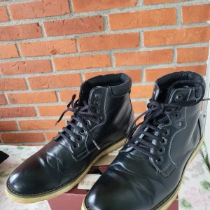 Svarta kängor från C5 i skinn - Svarta kängor från C5 i äkta skinn med snörning och kontrasterande ljus sula. Skorna har en klassisk design med rund tå och vadderad kant upptill för extra komfort. Perfekta för dig som vill ha en stilren och tidlös look.