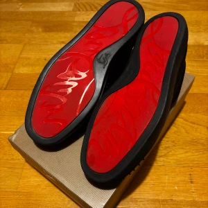 Christian Louboutin svarta med spikes  - Säljer ett par svarta sneakers från Christian Louboutin med ikoniska röda sulor och svarta nitar på tån. Skorna har snörning, rund tå och är tillverkade i mocka med detaljer i skinn. Kommer med originalkartong, dustbag och extra nitar. Helt nya men använda 1-2 gånger tyckte inte det var min stil. Om mer frågor komm pv!
