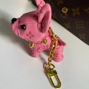 Vuitton väsktillbehör och nyckelringar - Säljer en klassisk  Vuittonväsktillbehör och nyckelringar i brunt med ikoniskt monogram och guldfärgade detaljer. har en kedja och tryckknapp framtill. Perfekt för dig som gillar lyxiga accessoarer och vill sticka ut med en snygg design.