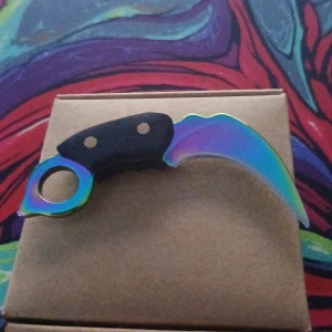 Karambit regnbågsfärgad kniv - Unik karambit-kniv med regnbågsfärgad böjd klinga och svart handtag. Perfekt för samlare eller cosplay. Kniven ser oanvänd ut och har inga synliga repor eller skador. Levereras i originalförpackning.