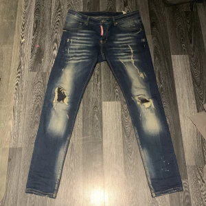 Dsquared2 blåa jeans - Säljer ett par blå jeans från Dsquared2 med coola slitningar och hål på knäna. Jeansen har en skinny passform, klassisk femficksdesign och snygga detaljer som blekta partier och färgstänk. Röd Dsquared2-logga vid gylfen. Perfekt för dig som gillar streetstyle.