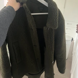 Olivgrön teddyjacka från COS, strl 36 - Mysig olivgrön teddyjacka från COS i storlek 36. Jackan har en klassisk krage, raka ärmar och stängs med knappar framtill. Tillverkad i ett fluffigt teddy-material som håller dig varm under kyliga dagar. Perfekt för dig som gillar en stilren och trendig look.