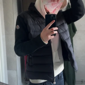 Moncler cardigan - Svart pufferjacka från Moncler med quiltad front och stickade ärmar. Jackan har dragkedja, hög krage och röda samt mörkblå detaljer längs sidorna med Moncler-logga. Materialet är dunfyllning och stickad polyester, perfekt för kyliga dagar.