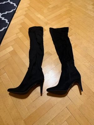 Svarta höga stövlar med spetsig tå - Säljer ett par svarta höga stövlar med spetsig tå och klack. Stövlarna har dragkedja på sidan och är gjorda i mocka. Snygg och stilren modell som passar till många olika outfits. Perfekt för dig som gillar att sticka ut med höga boots. Helt nya. Prislapp kvar. 