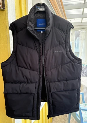 Svart dunväst från Jack & Jones - Svart dunväst från Jack & Jones Originals Studio med quiltad design och hög krage. Västen har två stora fickor framtill och stängs med dragkedja. Perfekt för lager-på-lager och har en stilren look som funkar till många outfits. Är stor i storleken. 