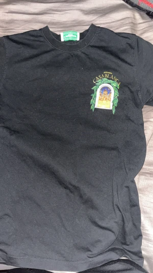 Svart Casablanca t-shirt med tryck - Svart t-shirt från Casablanca med färgstarkt tryck av en byggnad och gröna blad på både bröstet och ryggen. Rund hals och korta ärmar. Trycket har detaljer i gult, blått, orange och grönt. Perfekt för dig som gillar unika motiv och streetwear.