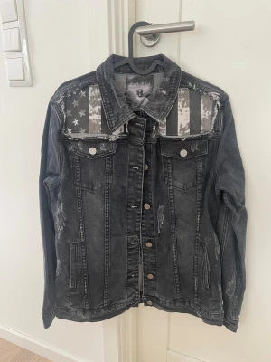 Svart jeansjacka Rock Rebel med flaggdetalj - Svart jeansjacka från Rock Rebel med slitna detaljer och stora svartvita flaggpatchar fram och bak. Jackan har knappar framtill, två bröstfickor med lock och långa ärmar. Snyggt broderad logga på ryggen och coolt, edgy uttryck.