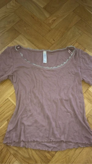 Rosa ribbad topp med spets från Lindex - Supersöt ljusrosa ribbad topp från Lindex i storlek XS. Toppen har korta ärmar, rundad halsringning med grå spetsdetalj och vågig kant nedtill. Perfekt för dig som gillar romantiska och mjuka vibes.