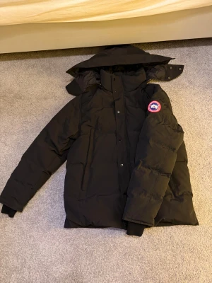 Canada Goose jacka  - Säljer en svart dunjacka från Canada Goose med klassisk patch på ärmen. Jackan har hög krage, avtagbar huva och stängs med både dragkedja och knappar. Perfekt för kalla vinterdagar och riktigt snygg streetstil.