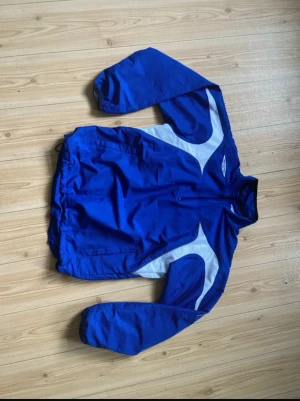 Blå  Umbro vindjacka - Säljer en blå och vit vindjacka från Umbro med sportig design. Jackan har meshfoder, snörning i midjan och dragkedja framtill. Perfekt för dig som gillar retrostil och vill sticka ut. Klassisk logga på bröstet och ryggen. Den är S för man, passar som L på kvinnor