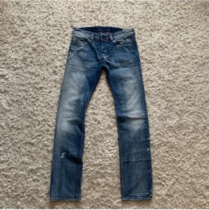 Diesel Safado jeans blå W30 L34 - Säljer ett par snygga Diesel Safado jeans i tvättad blå färg med slitningar och coola detaljer. Klassisk femficksmodell med raka ben och normal passform. Jeansen har en schysst faded look och är tillverkade i slitstarkt denim. Pris kan diskuteras vid snabb affär.