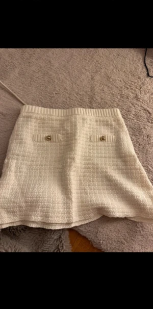 Vit kortkjol med fickor och knappar - Säljer en vit kortkjol med snygg struktur och två dekorativa fickor framtill med guldfärgade knappar. Kjolen har en ribbad midja och är tillverkad i ett tjockare, stickat material som ger en exklusiv känsla.