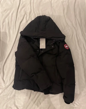 Canada Goose Macmillan  - Säljer en svart dunjacka från Canada Goose med huva och klassisk logga på ärmen. Jackan har dragkedja och knappar framtill, samt praktiska fickor. Insidan har en axelrem med Canada Goose-tryck. Perfekt för kalla vinterdagar. Helt ny aldrig använd. 