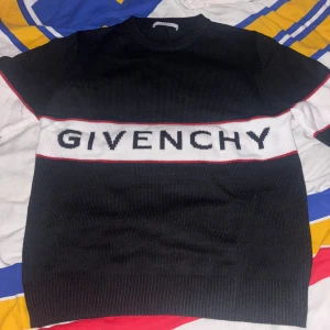 Svart stickad tröja från Givenchy - Snygg svart stickad tröja från Givenchy med vit panel och logga över bröstet. Tröjan har röda detaljer längs panelen och vita partier på ärmarna. Rund hals och långa ärmar med ribbade muddar. Perfekt för dig som gillar streetstyle och designer. Tröjan är i storlek L men passar även M. Priset kan diskuteras!!!