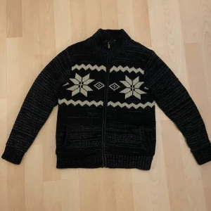 Svart kofta från Livergy - Svart stickad kofta med dragkedja. Framsidan har ett snyggt vinterinspirerat mönster med stora vita stjärnor och zigzag-ränder. Koftan har två fickor framtill och långärmad design, perfekt för kyliga dagar. i mycket gott skick, inga defekter