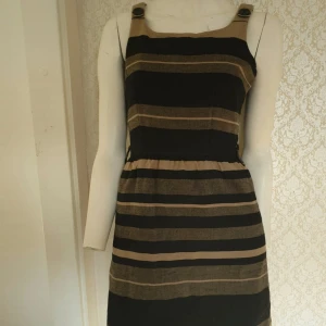 Randig klänning med knappar - Snygg randig klänning i svart och beige med knappar på axelbanden. Perfekt för en stilren look. Klänningen har en normal passform och är knälång.