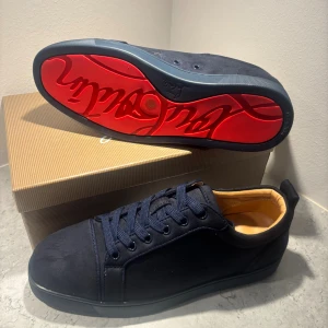Christian Louboutin marinblå sneakers - Exklusiva marinblå sneakers från Christian Louboutin i mocka med klassisk röd sula och snygga snördetaljer. Skorna har rund tå, platt sula och lyxig beige innersida i skinn. Perfekta för dig som vill sticka ut med en ikonisk design och vara stilig. Kan sänkas lite vid snabbaffär