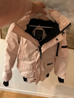 Ljusrosa Canada Goose dunjacka - Säljer en ljusrosa Canada Goose dunjacka med huva och äkta pälsdetalj. Jackan har flera fickor, svart foder och klassisk logga på ärmen. Dragkedja framtill och ribbade muddar vid ärmslut. Perfekt för kalla vinterdagar.xxs len passar cxs-s