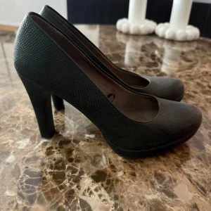 Svarta pumps med strukturmönster från Tamaris - Snygga svarta pumps från Tamaris med rund tå och hög klack. Skorna har en diskret struktur som ger en cool reptil-look och är perfekta för att lyfta vilken outfit som helst. Klassisk modell med platt sula framtill och stilren design.