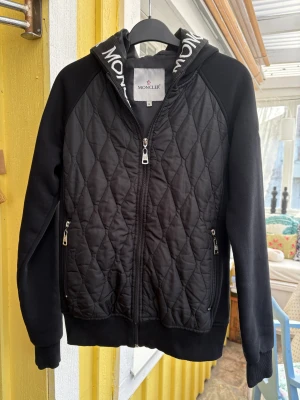 Svart quiltad jacka från Moncler - Svart quiltad jacka från Moncler med huva och broderad logga på ärmen. Jackan har dragkedja framtill, två fickor med dragkedja och ribbade muddar. Snyggt mönstrad med Moncler-tryck på huvan och diamantquiltad front och rygg. 