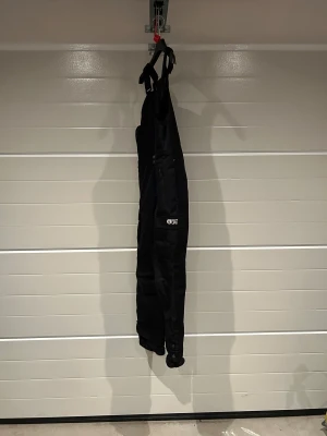 Svarta täckbyxor från Picture Organic - Svarta junior täckbyxor från Picture Organic med hängslen och dragkedja framtill. Byxorna har flera fickor med dragkedja och knappstängning, samt förstärkta benslut. Grymma skidkläder som jag även säljer jacka till. Fodrade och med smarta fickor, snölås m.m storlek 14, motsvara xs. 