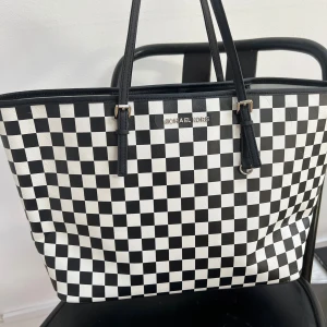 Svartvit rutig shoppingväska Michael Kors - Snygg shoppingväska från Michael Kors med svartvitt rutigt mönster och silverfärgade detaljer. Väskan har två långa handtag, insida med dragkedja och är tillverkad i skinn. Perfekt för dig som gillar statement-accessoarer och vill sticka ut.