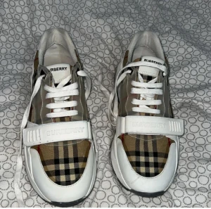 Burberry sneakers med klassiskt rutmönster - Säljer ett par snygga Burberry sneakers med det ikoniska beige, svarta och röda rutmönstret. Skorna har vita detaljer i skinn, snörning och en bred rem med Burberry-logga över foten. Perfekta för dig som vill sticka ut med en lyxig och trendig look.