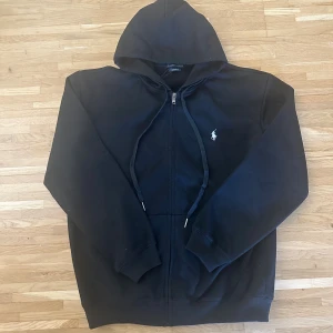 Svart hoodie från Ralph Lauren - Svart hoodie från Ralph Lauren med dragkedja och huva. Klassisk design med vit broderad logga på bröstet och snörning i huvan. Tillverkad i mjuk bomull, perfekt för chill dagar. Långa ärmar och ribbade muddar för extra komfort.