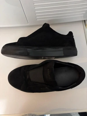 Svarta sneakers Axel Arigato mocka - Snygga svarta sneakers från Axel Arigato i mjuk mocka med platt sula och rund tå. Diskret guldtext på sidan och elastisk panel över vristen för enkel påtagning. Kommer med original dustbag. Perfekt för dig som gillar minimalistisk stil.