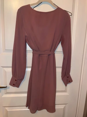 Rosa omlottklänning från Sisters Point - Supersnygg kort rosa omlottklänning från Sisters Point med långa ärmar, knytskärp i midjan och breda manschetter med knappar. Klänningen har en v-ringad omlottfront och är tillverkad i ett lätt, flowy material som ger en feminin siluett.