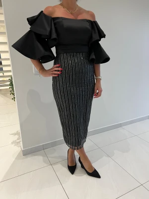 Svart offshoulder klänning med volang - Supersnygg svart offshoulder klänning med stora volangärmar och figurnära passform. Kjolen är glittrig med vertikala silverränder och slits bak. Klänningen har dragkedja i ryggen och är perfekt för dig som vill sticka ut med en elegant och trendig look.