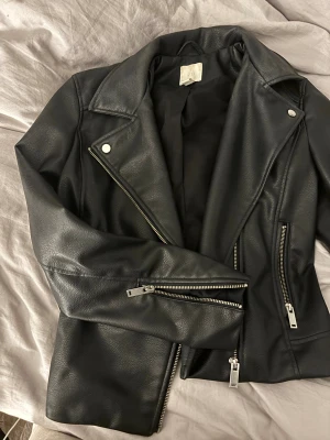 Svart skinnjacka från H&M med dragkedjor - Snygg svart skinnjacka från H&M med klassisk bikerstil. Jackan har silvriga dragkedjor framtill och på ärmarna, bred krage och coola detaljer i metall. Perfekt för dig som vill ha en edgy look och gillar tidlös stil.