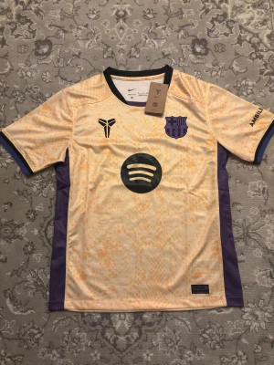Barcelona x Kobe 25/26  - Unik Barcelona x Kobe fotbollströja från Nike i Dri-Fit material. Tröjan är ljusgul med lila detaljer och har ett subtilt camouflage-mönster. FCB-logga i lila, Kobe-symbol och Spotify-logga på bröstet. Svart krage och lila paneler på sidorna. Perfekt för dig som vill sticka ut på planen.
