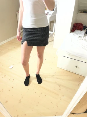 Svart kortkjol med slits från H&M Divided - Säljer en svart kortkjol från H&M Divided med vita tunna ränder och en liten slits framtill. Kjolen har en broderad hjärtdetalj i ljusrosa på linningen och stängs med dragkedja i sidan. Snygg och trendig modell som passar perfekt till sneakers eller boots. 