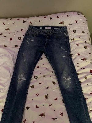 Replay MICKYM blå jeans slim tapered - Säljer ett par blå Replay MICKYM jeans med slim tapered fit. Jeansen har slitna detaljer och snygga slitningar på benen, klassisk femficksdesign och knappgylf. Materialet är jeans med en skön stretch. Passar dig som gillar en modern och smal siluett.