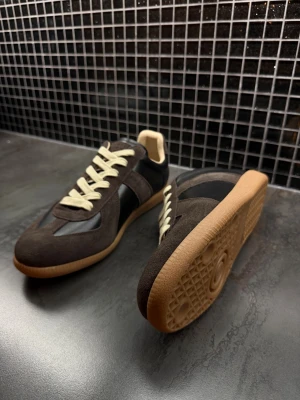 Maison margiela gats  - Snygga sneakers i mörkbrun mocka och svart skinn med beige snörning och gummisula. Klassisk retrostil med rund tå och platt sula, perfekta för dig som gillar stilrena och tidlösa skor. Skorna har en låg profil och kontrasterande detaljer.