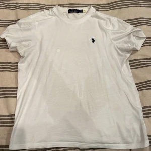 Vit t-shirt från Polo Ralph Lauren - Vit t-shirt från Polo Ralph Lauren med klassisk rund hals och korta ärmar. T-shirten har det ikoniska blå Polo-logot broderat på bröstet. Tillverkad i mjuk bomull för en skön känsla och clean look. Finns en fläck som jag inte orkat ta bort då tröjan har blivit för liten.