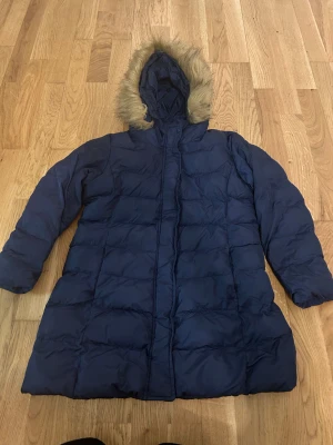 Mörkblå lång dunjacka från Uniqlo - Säljer en mörkblå lång dunjacka från Uniqlo med huva och fuskpälsdetalj. Jackan är quiltad och har dragkedja framtill. Perfekt för kalla dagar och riktigt skön att bära. Klassisk och stilren design som håller dig varm hela vintern.